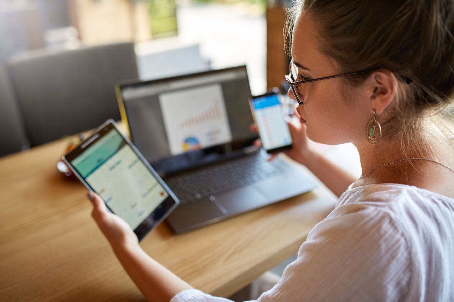 Woman holding phone in one hand, tablet in another, at a laptop computer.