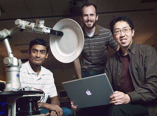 Three men posed around a robotic arm that is holding a plate