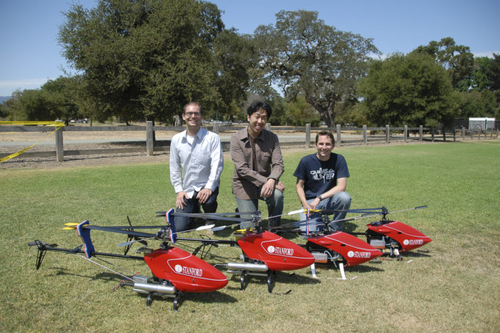 Three men kneeling behind three radio-controlled helicopters outside