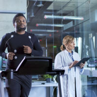 Male Athlete Walks on a Treadmill with Electrodes Attached to His Body while Sport Scientist Interacts with Touchscreen and Supervises EKG Status. In the Background Laboratory with High-Tech Equipment.