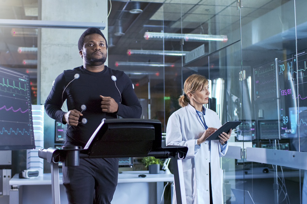 Male Athlete Walks on a Treadmill with Electrodes Attached to His Body while Sport Scientist Interacts with Touchscreen and Supervises EKG Status. In the Background Laboratory with High-Tech Equipment.