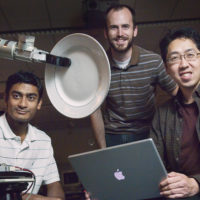 three men posed around a robotic arm holding a plate