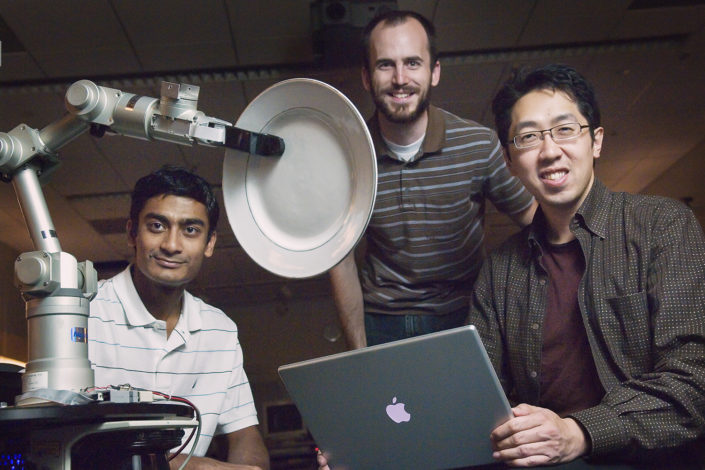 three men posed around a robotic arm holding a plate