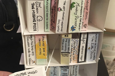 A paper bookshelf with cards made to look like books. A person is pulling on the Misérables card to show how it makes a sign reading "D-Library Favorites for Hugo" pop up.