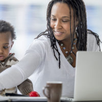 A woman of African descent and her baby boy are at home. The mother is working on a laptop computer. She is being distracted by her son, who was playing with a pencil.