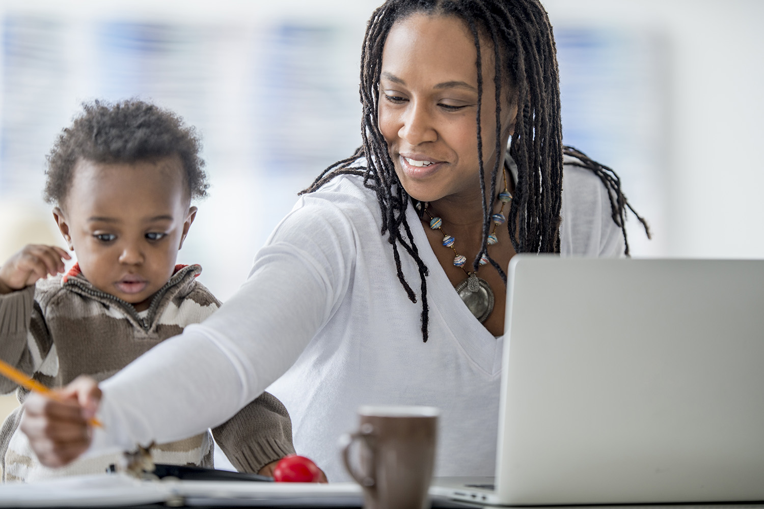 A woman of African descent and her baby boy are at home. The mother is working on a laptop computer. She is being distracted by her son, who was playing with a pencil.