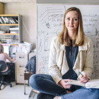 A young woman holds a notebook as she sits on a table in a lab.