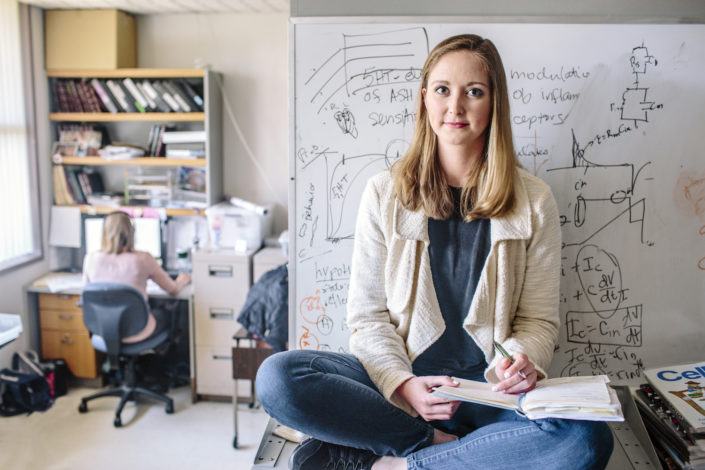 A young woman holds a notebook as she sits on a table in a lab.