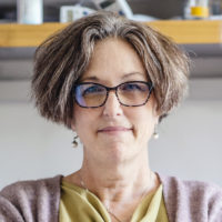 A woman with short hair and glasses stands in a lab and looks at the camera.