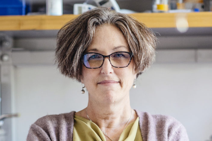 A woman with short hair and glasses stands in a lab and looks at the camera.