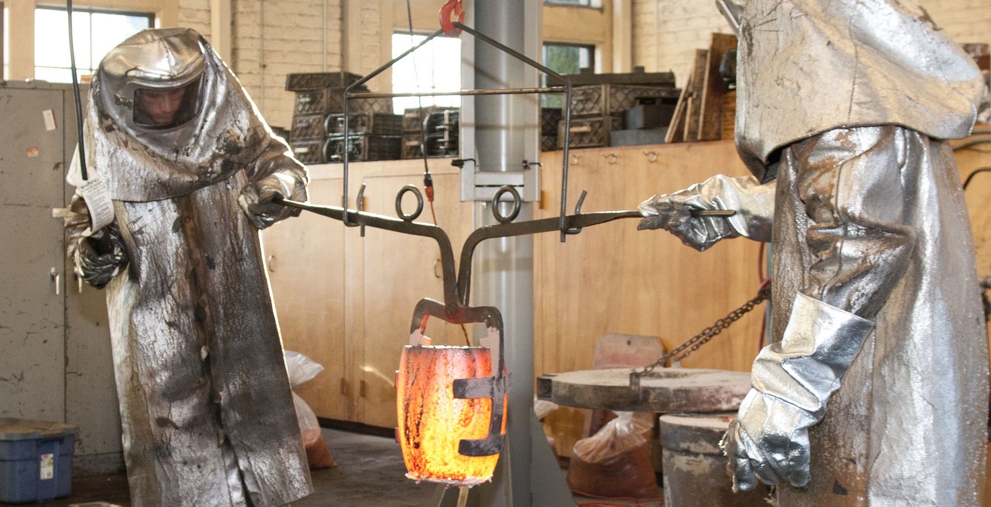 Two people in full-body protective gear moving a container of molten metal