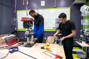 One person is cutting a dowel with a hacksaw while another watches