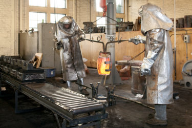 Two people in full-body protective gear moving a container of molten metal toward molds