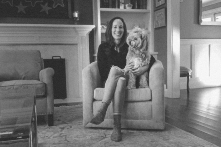 Amy Zegart smiling, sitting in a chair with her dog, whose head is cocked to the side