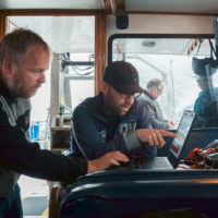 Two men inside a boat, looking at a computer screen