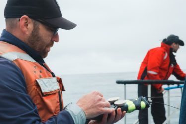 Person in foreground holding a device with another person in the background on the pulpit of a separate boat