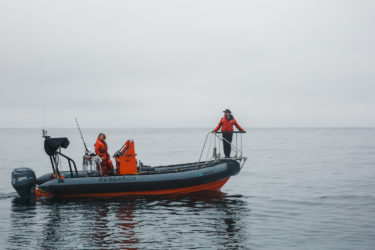 Two people standing on a boat, one near the steering wheel and one on a pulpit at the front