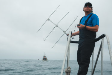 Man standing on pulpit of boat with an antenna in his hands. A large boat in the background.