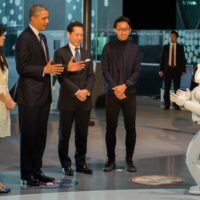 Four people facing a human-like robot. One person is Barack Obama. Obama and the robot have their hands raised in similar ways.