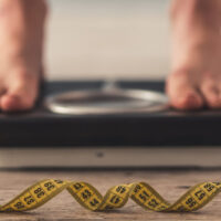 Cropped image of woman feet standing on weigh scales, on gray background. A tape measure in the foreground